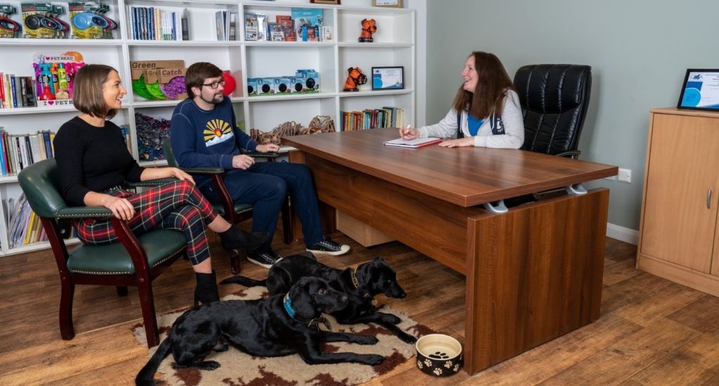 A professional behaviourist at the Company of Animals Pet Centre taking notes during a consultation with a couple. Two black dogs lie calmly on the floor, illustrating a safe, controlled environment for addressing the root causes of biting.